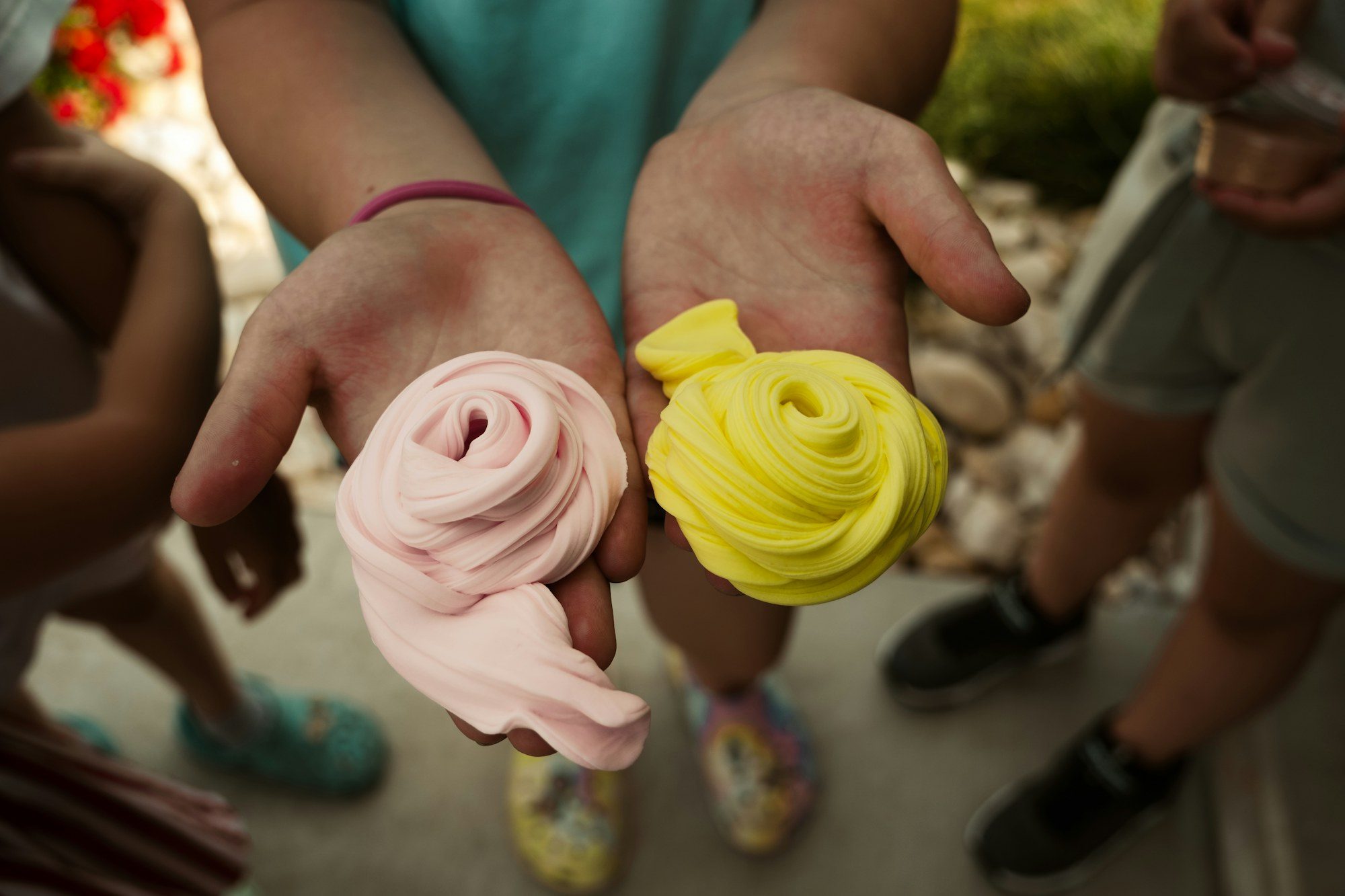 Two open hands holding a coil of pink dough and a coil of yellow dough, with other children standing out of focus behind.