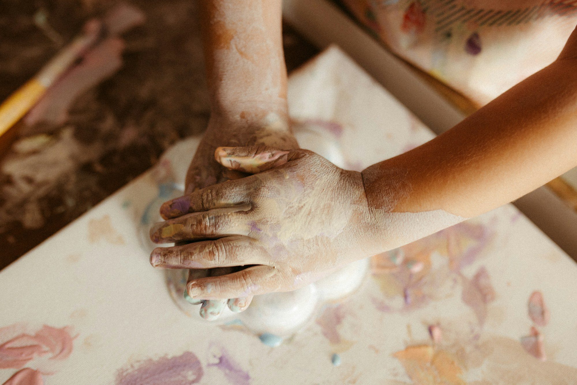 A young child's hands, covered in pale paint, pressed flat on a canvas already covered in smeared color.
